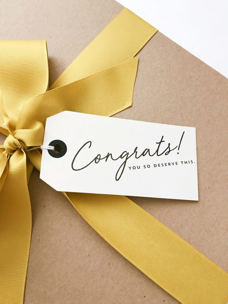 a close up of a gift box wrapped in classic brown paper and featuring a gold ribbon, a white gift tags reads 'congrats, you so deserve this!'