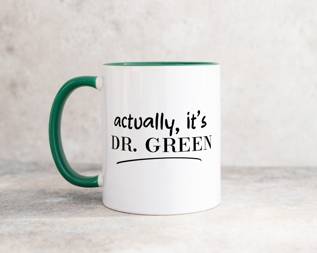 a classic white mug with a dark green handle, pictured in front of a grey background. the mug reads 'actually, it's DR Green'.