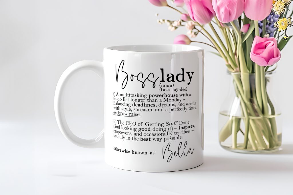 a classic white standard mug sat in front of a clear glass vase holding pink flowers. The writing on the mug reads 'Boss Lady' followed by a dictionary-style definition and a personalised name.