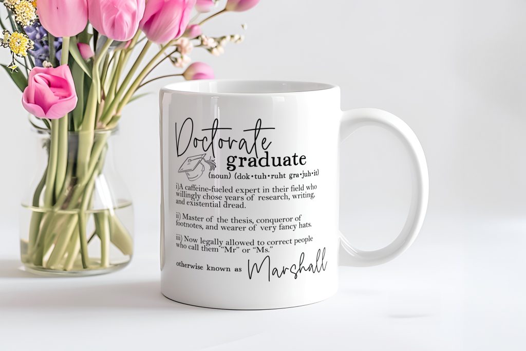 a white mug pictured in front of a vase of pink tulips - the mug features the phrase 'doctorate graduate' followed by a funny description and a personalised name.