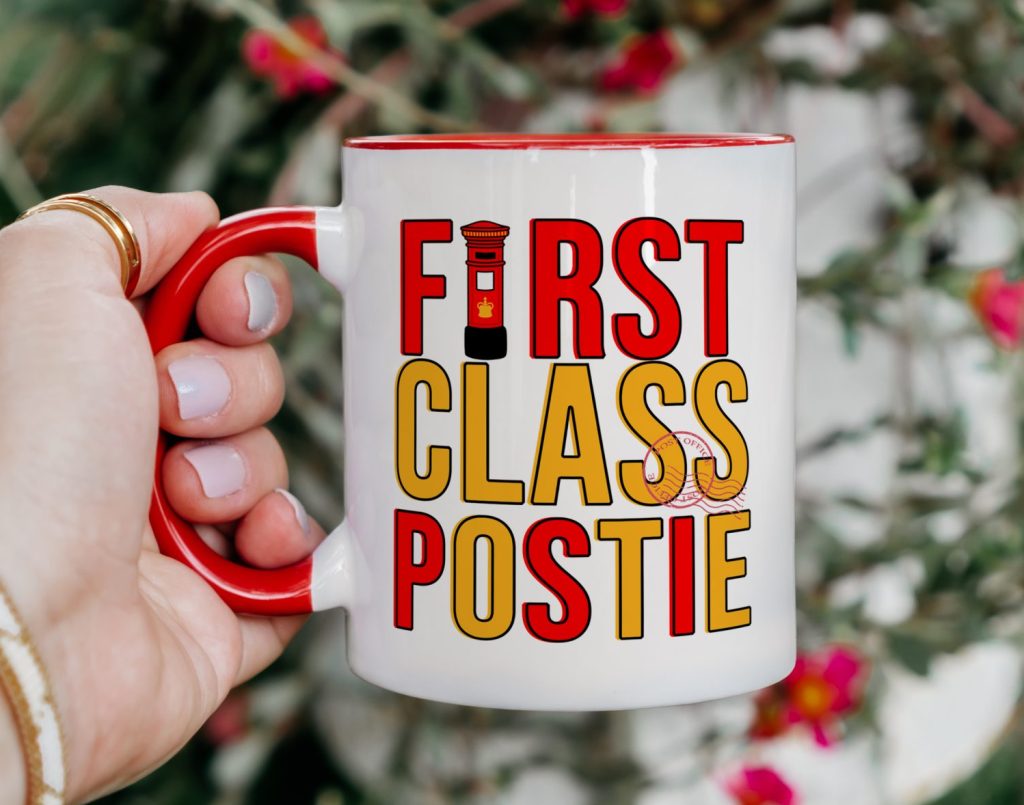 a white classic ceramic mug with a red handle, showcasing the phrase 'first class postie', being held in front of a shrub plant background.
