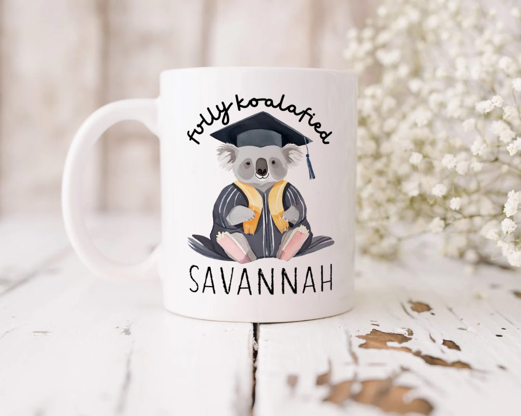 a white ceramic mug pictured in front of a distressed-white-wooden background, the mug design features a koala wearing a graduation gown and the phrase 'fully koalafied' alongside a personalised name.