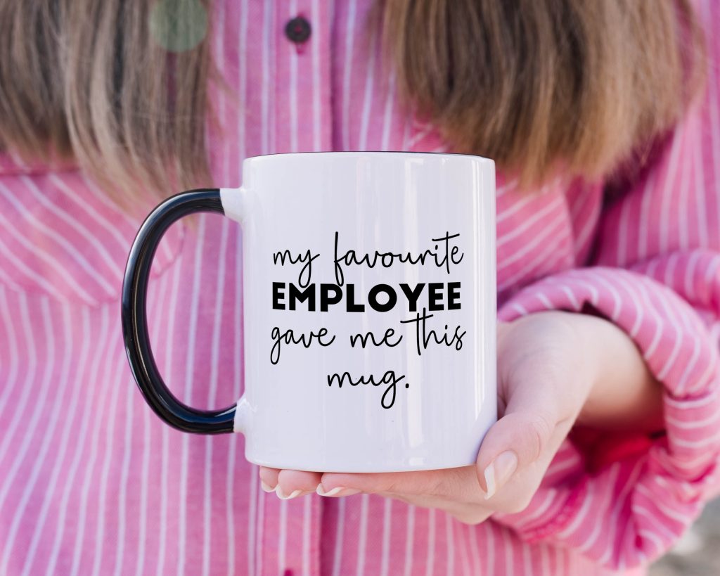a classic white mug featuring a black handle, being held against a pink shirt background, with the phrase 'my favourite employee gave me this mug'.