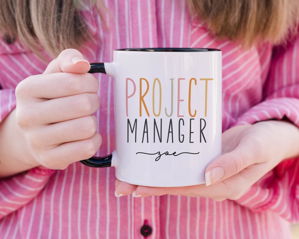 a classic white mug with a black handle, the mug is being held by a person wearing a pink and white striped shirt - the mug features the words 'project manager' followed by a personalised name.